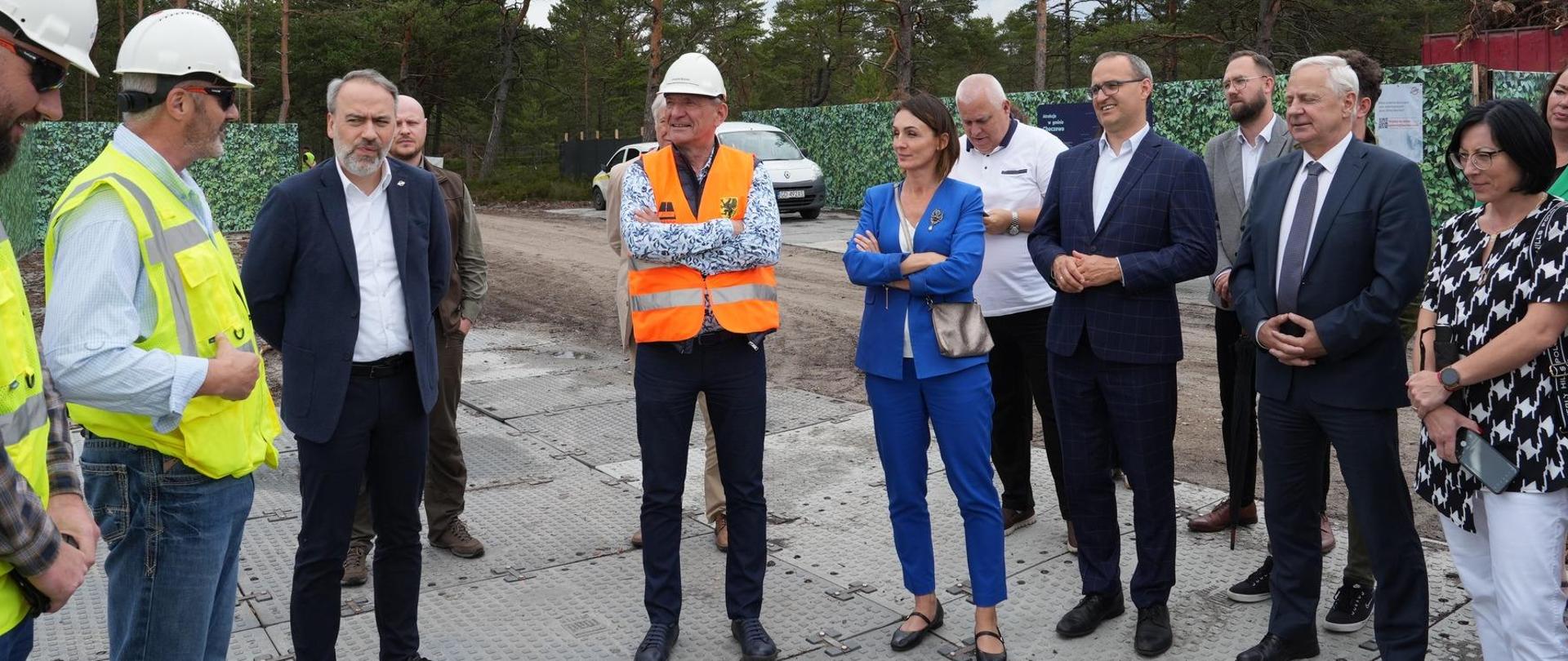 grupa osób stoi na placu, wśród nich są osoby w kaskach budowlanych