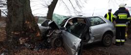 Zdjęcie przedstawia samochód marki Audi po uderzeniu w drzewo oraz strażaków i policjanta