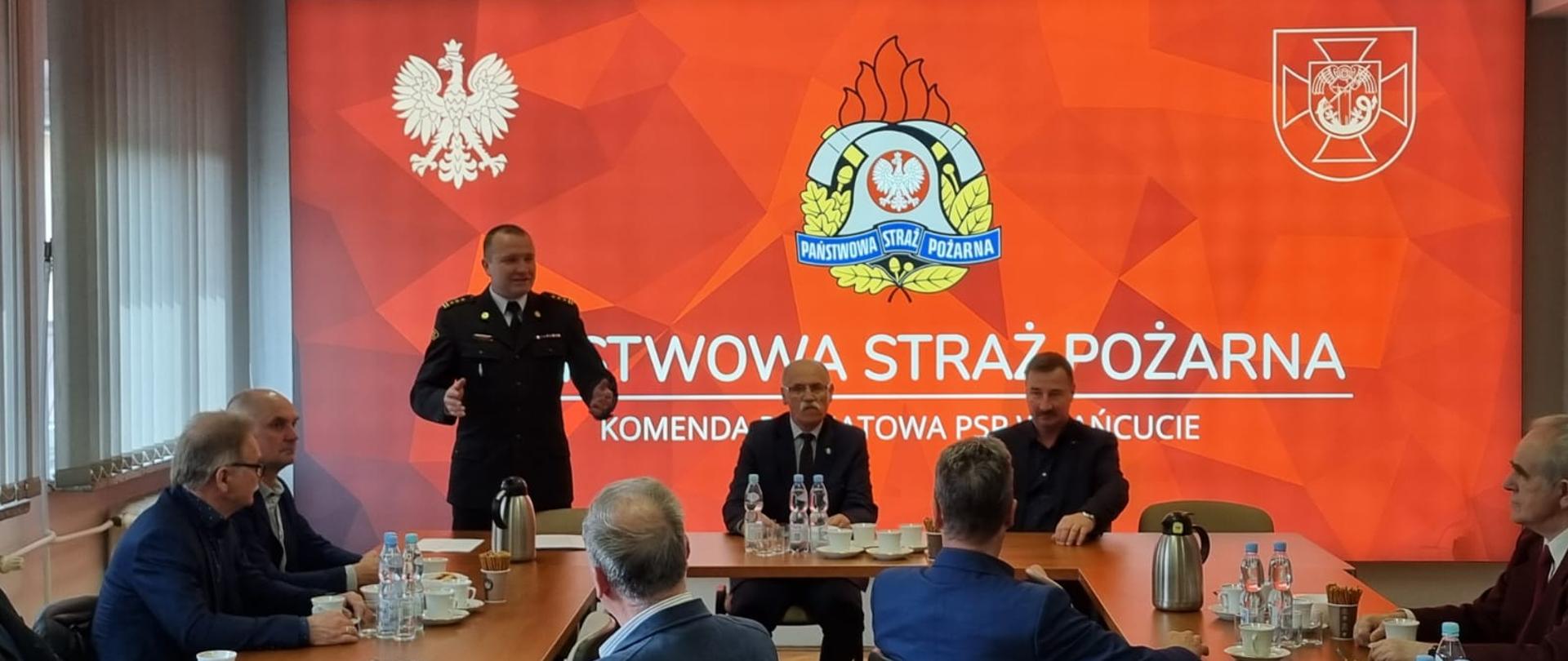 Zdjęcie przedstawia świetlicę Komendy Powiatowej. Na pierwszym planie przy stole konferencyjnym od lewej stojący i przemawiający Komendant Powiatowy Państwowej Straży Pożarnej w Łańcucie starszy brygadier Bogusław Golenia, następnie siedzą Prezes Zarządu Oddziału Wojewódzkiego Związku Emerytów i Rencistów Pożarnictwa w Rzeszowie Tadeusz Cyrek oraz Prezes Zarządu Oddziału Powiatowego Związku Emerytów i Rencistów Pożarnictwa w Łańcucie Janusz Cierpisz. W tle podświetlony logotyp prezentujący godło Polski, logo powiatu łańcuckiego oraz po środku logo państwowej straży pożarnej, pod nim napis "Państwowa Straż Pożarna Komenda Powiatowa PSP w Łańcucie"