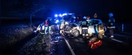 Zdjęcie przedstawia strażaków podczas udzielania pomocy ofiarom wypadku w tle rozbite pojazdy.