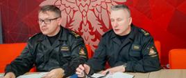 Zdjęcie przedstawia dwóch strażaków podczas wideokonferencji kadry kierowniczej PSP woj. mazowieckiego w sprawie wsparcia Narodowego Programu Szczepień. W tle ścianka koloru czerwonego z godłem RP.
