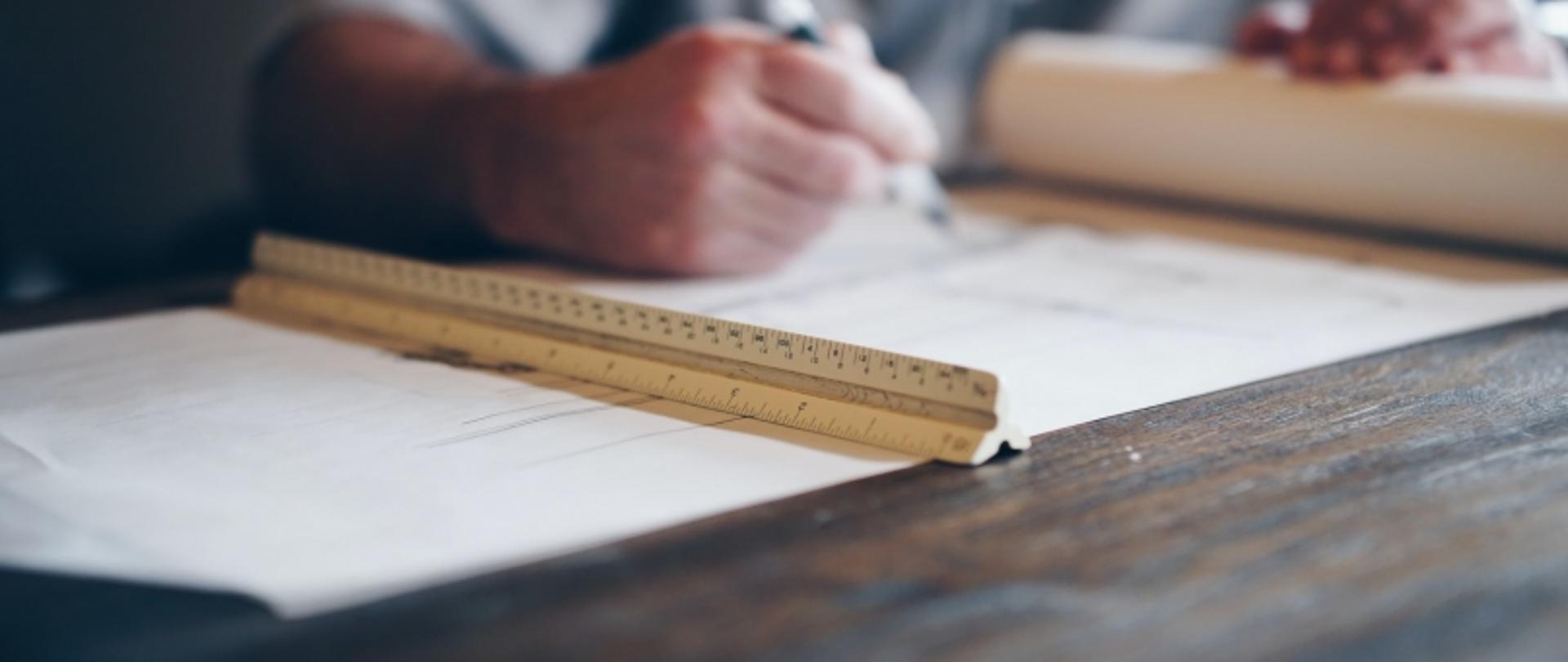 Praca nad rysunkiem technicznym przy stole z papierem, linijką i długopisem.