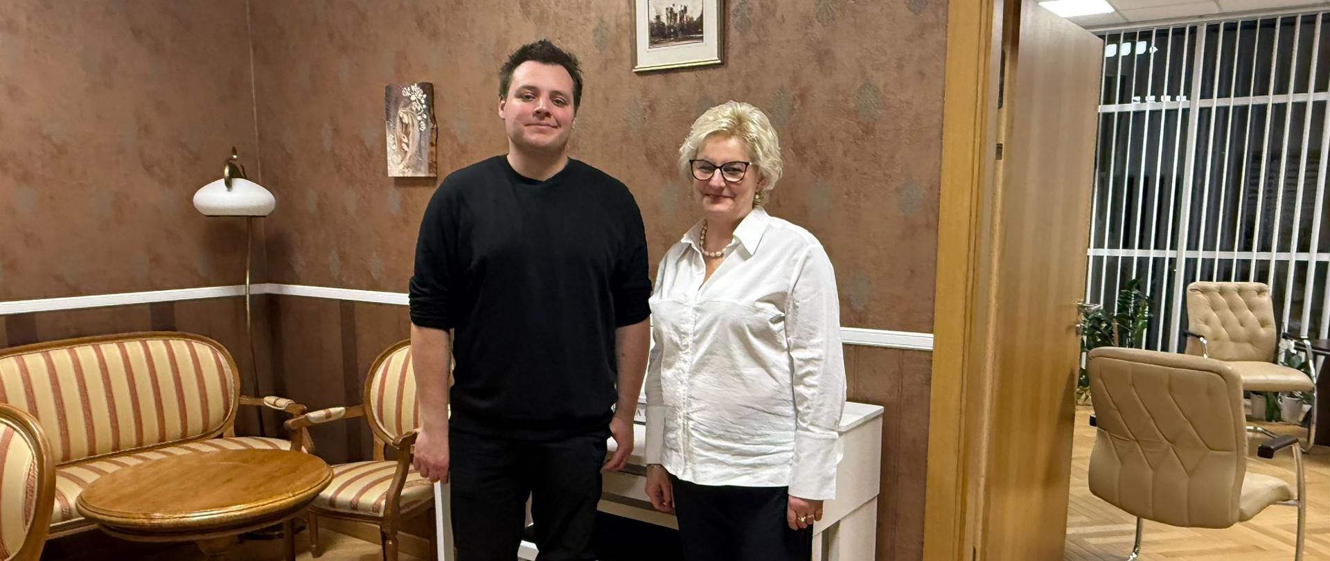 Dwie osoby stoją w eleganckim pomieszczeniu z drewnianą podłogą, brązowymi ścianami i klasycznymi krzesłami. Po lewej stronie stoi mężczyzna w czarnym swetrze i czarnych spodniach, a po prawej kobieta w czarnych spodniach i białej koszuli. W tle widoczny jest biały pianino oraz obrazy na ścianie.