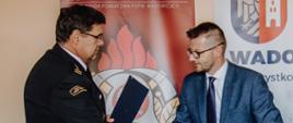 Widok z boku. Komendant Powiatowy podaje dłoń Burmistrzowi Wadowic na tle banerów KP PSP w Wadowicach oraz Gminy Wadowice. 