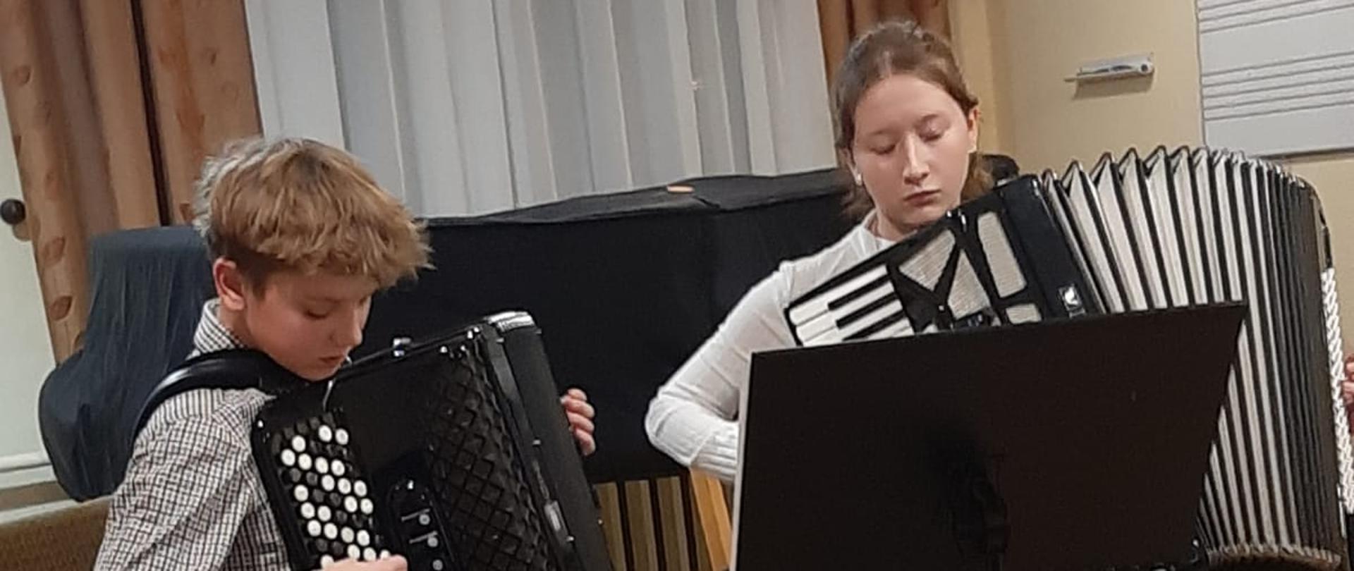 Zdjęcie w sali. Na pierwszym planie duet akordeonowy. Uczennica i uczeń grają na akordeonie siedząc. Przed nimi pulpit z nutami. Za uczniami fortepian przykryty pokrowcem, obok ława fortepianowa. Za fortepianem okno przykryte firanka i dwoma zasłonami w listki. Na ścianie po prawej stronie biała tablica, na której narysowane nutki.