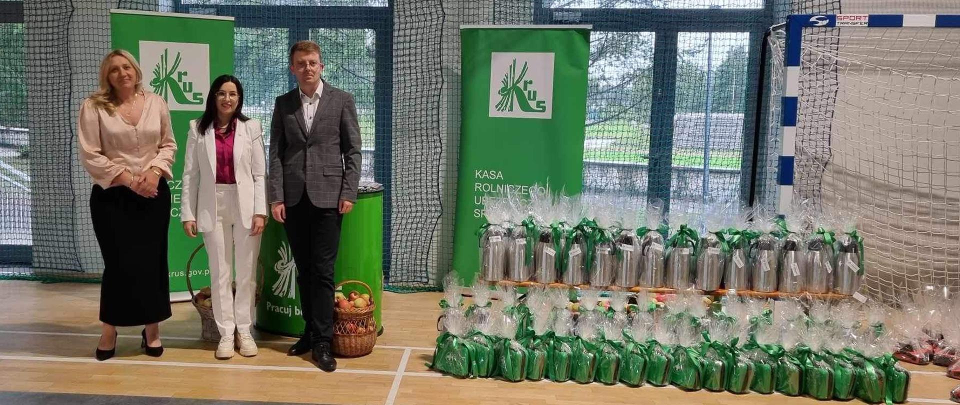 Na tle stoiska informacyjnego KRUS stoją dwie kobiety i jeden mężczyzna obok ozdobnie zapakowane nagrody