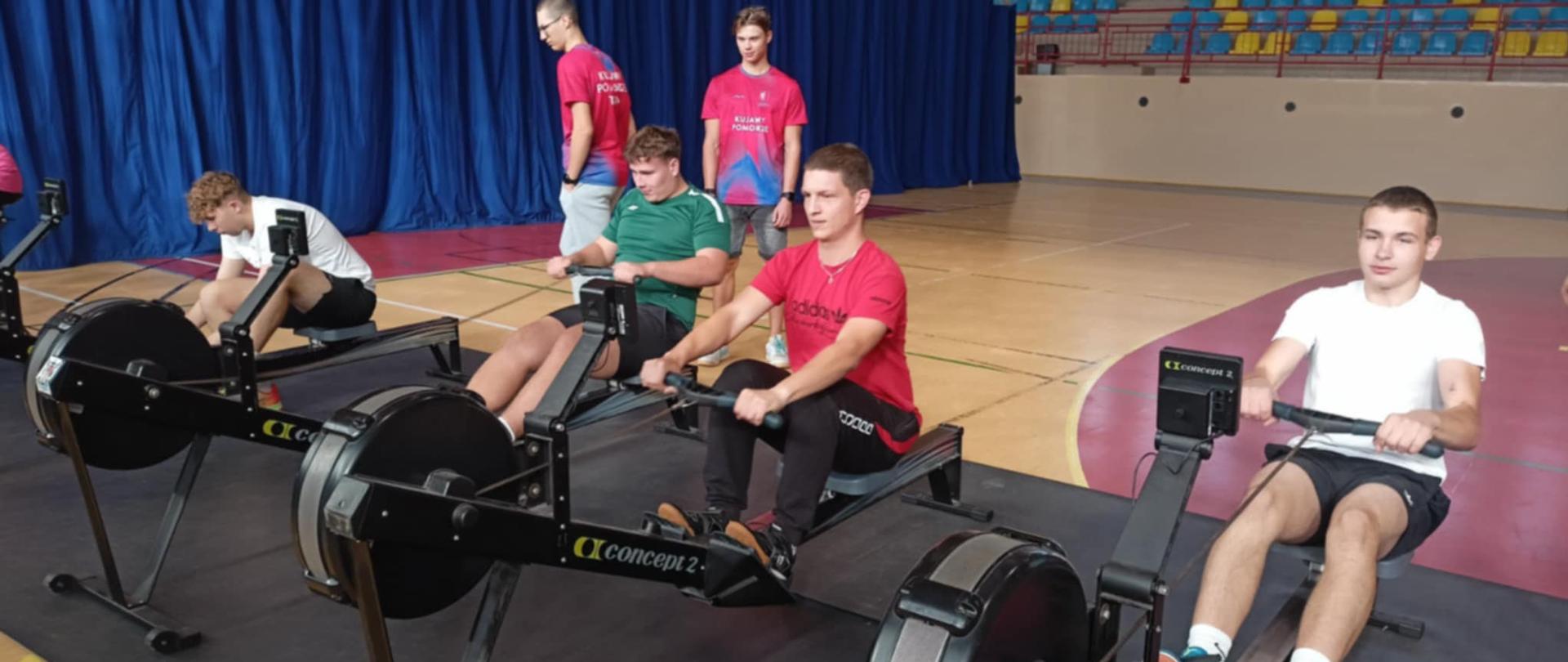 Na zdjęciu widać grupę młodych osób ćwiczących w sali gimnastycznej na ergometrach wioślarskich marki Concept2. W pierwszym planie, na czarnej macie ustawione są cztery wiosła treningowe w jednym rzędzie. Na każdym z nich siedzi jedna osoba i energicznie wykonuje ruchy wiosłowania.