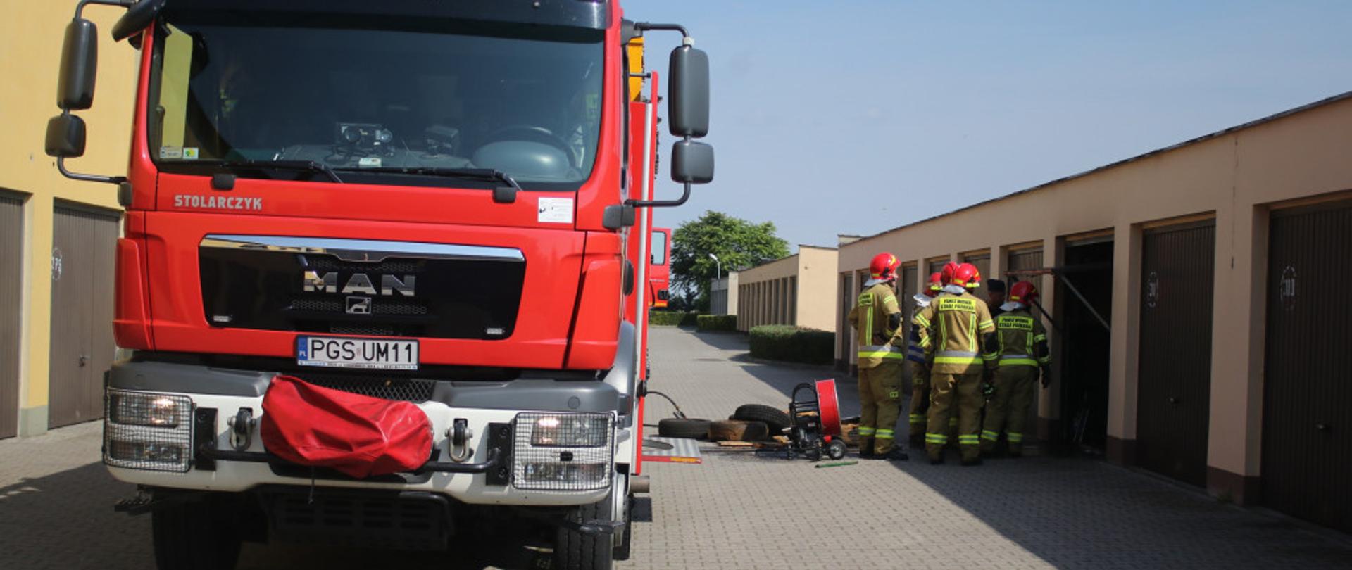 Zdjęcie przedstawia samochód pożarniczy. Obok znajduje się kilku strażaków, którzy prowadzą działania przy pożarze garażu.