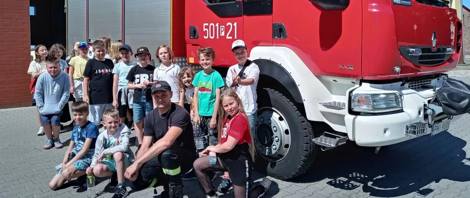 Na zdjęciu widzimy strażaka oraz dzieci. W tle widać samochód pożarniczy.