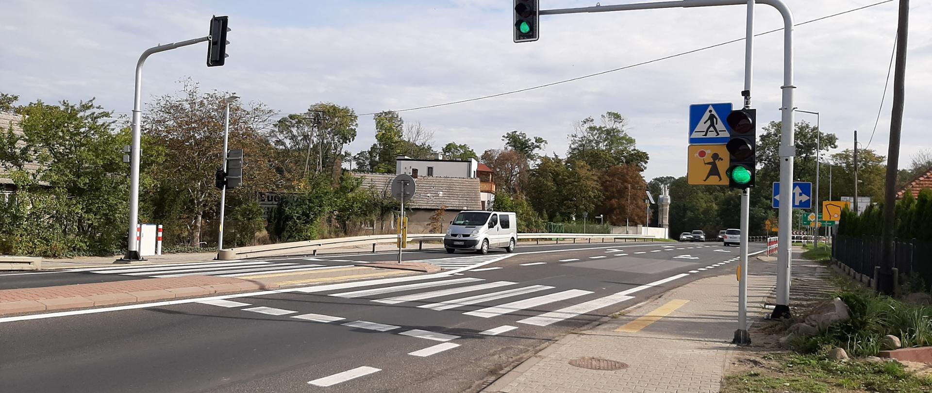 zdjęcie przedstawia przejście dla pieszych z sygnalizacją świetlną. W tle widoczne zabudowania.
