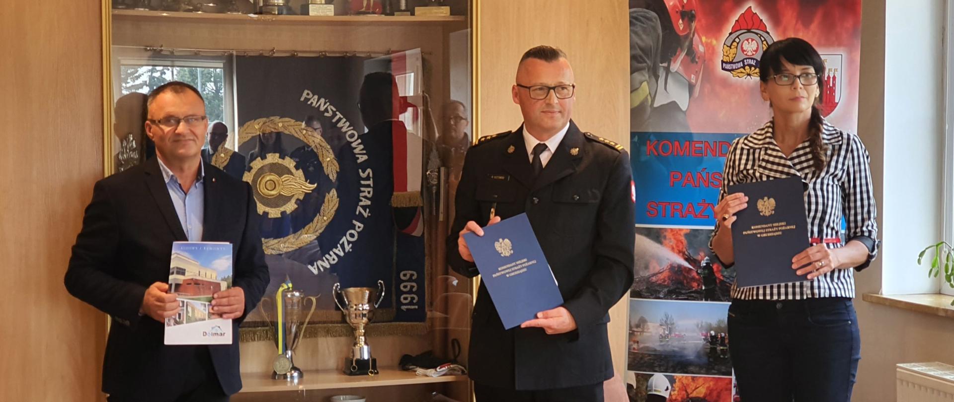 Zdjęcie przedstawia dwóch mężczyzn i jedną kobietę, którzy stoją i prezentują teczki z podpisanymi dokumentami. W tle plakat ze zdjęciami z akcji strażackich oraz witryna ze sztandarem i pucharami.