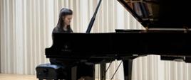 Dziewczyna brunetka gra recital fortepianowy na dużym czarnym fortepianie koncertowym na scenie sali koncertowej. Pianistka ubrana jest na czarno i siedzi przy instrumencie ustawionym na jasnej drewnianej podłodze, na tle jasnych pionowych paneli akustycznych. Na pierwszym planie widoczna jest publiczność siedząca w rzędach sali koncertowej i słuchająca występu w ramach Gdańskiego Festiwalu Muzycznego.