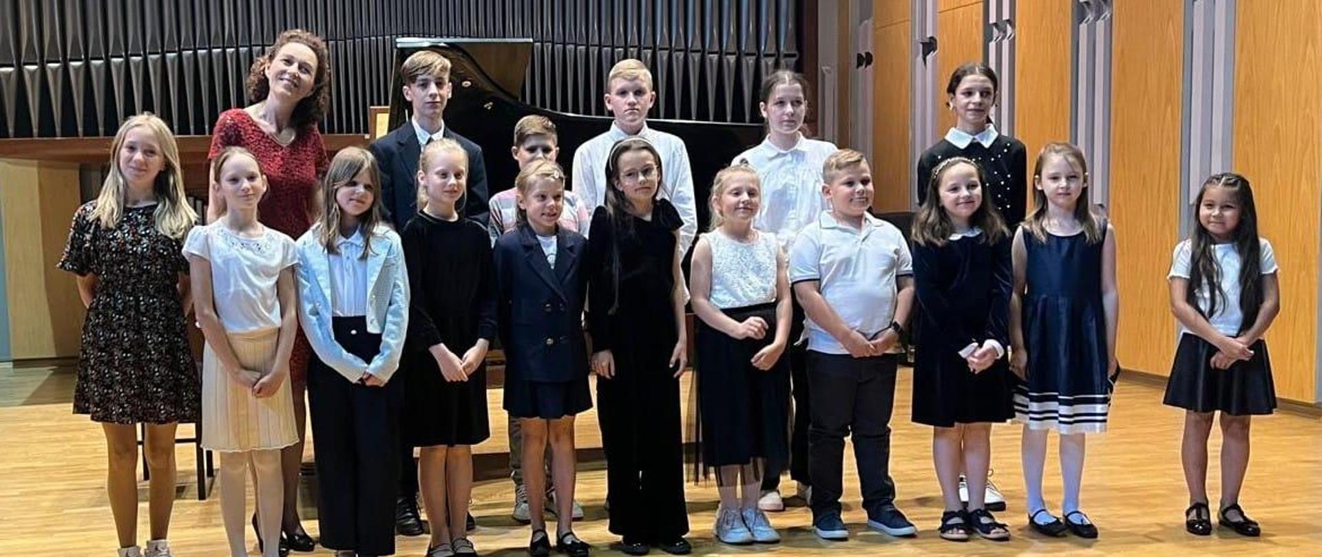 Wszyscy uczestnicy oraz nauczycielka organizująca koncert stoją w dwóch rzędach na scenie sali koncertowej. Za nimi organy