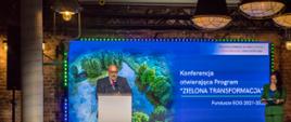 Kadr przedstawia zbliżenie na ekran podczas konferencji otwierającej Program 'Zielona Transformacja'. Za mównicą na scenie mężczyzna - Ambasador Islandii - Friðrik Jónsson.