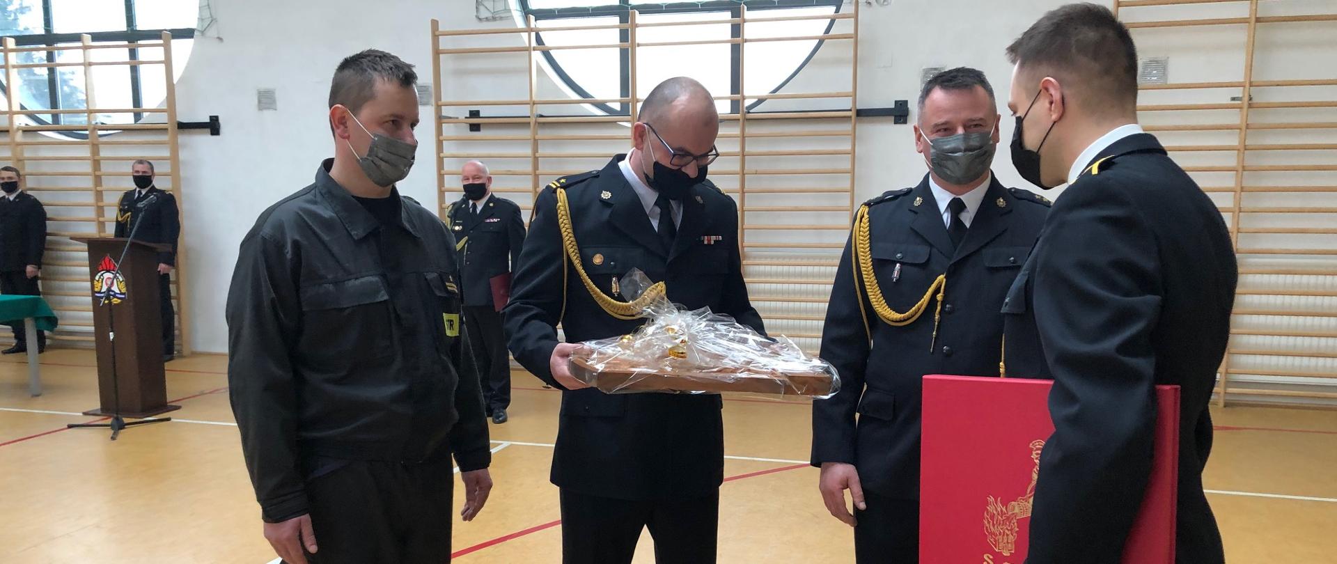 Delegacja strażaków dziękuje odchodzącemu ze służby. Jeden z nich trzyma w ręce upominek. Strażak odchodzący ma czerwone etui. W tle widać innych uczestników uroczystości..