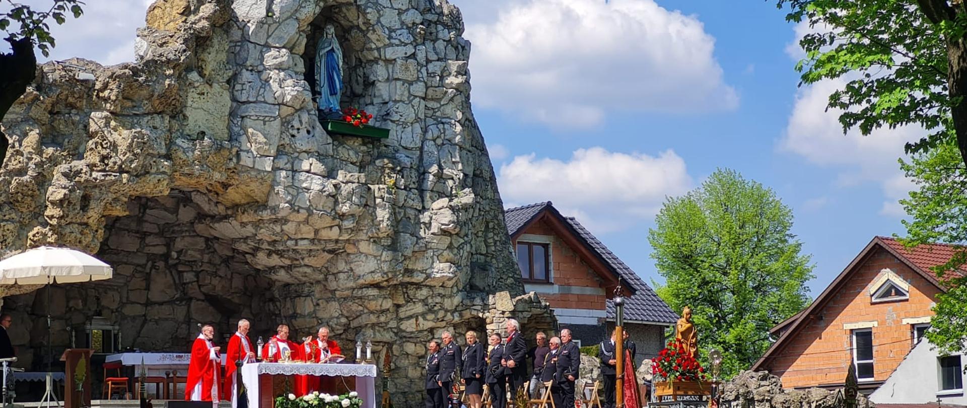 Na zdjęciu znajduje się ołtarz, księża, poczty sztandarowe oraz uczestnicy mszy świętej w grocie z kamieni na tle dachów z budynków mieszkalnych