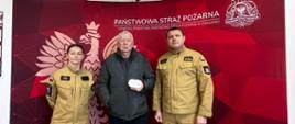 Trzy osoby pozują do zdjęcia przed ścianką Państwowej Straży Pożarnej. Pośrodku mężczyzna trzyma czujnik dymu, po jego bokach stoją dwaj strażacy w umundurowaniu służbowym.