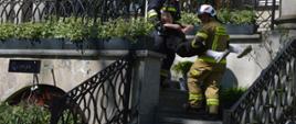 Ćwiczenia w Pakosławiu. Trwa akcja ratowniczo-gaśnicza. Strażacy schodami zewnętrznymi ewakuują kolejną osobę poszkodowaną. W tle budynek. 