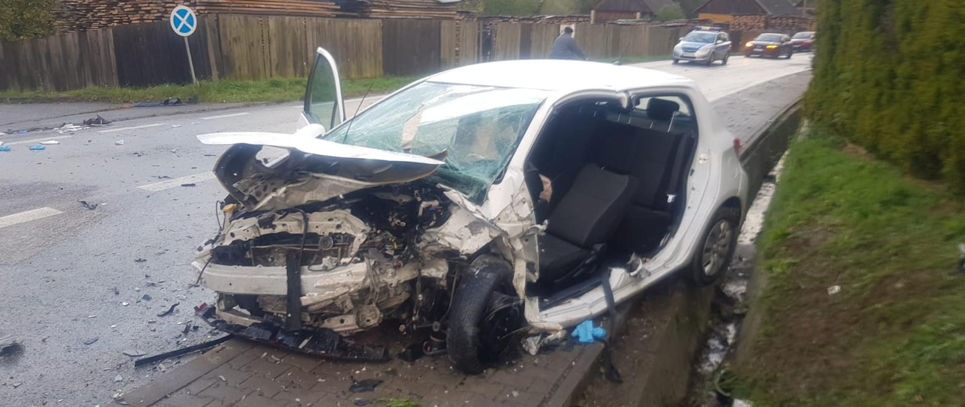 Zdjęcie przedstawia rozbity samochód osobowy po zderzeniu czołowym. Lewy bok został usunięty przez strażaków w celu wykonania dostępu do osoby poszkodowanej. W tle zabudowania miejscowości.