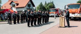 Szereg strażaków w ciemnych mundurach galowych i czapkach rogatywkach stoi na baczność podczas oficjalnej uroczystości na świeżym powietrzu.
