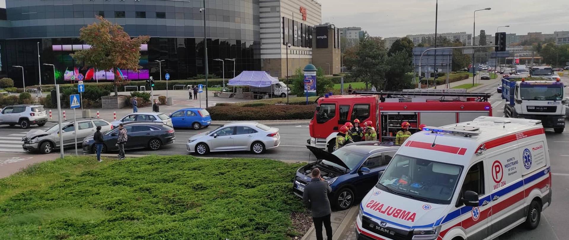 Na zdjęciu widoczne są dwa pojazdy, które brały udział w wypadku, pracujący strażacy oraz karetka pogotowia, straż pożarna. W tle budynek galerii handlowej.
