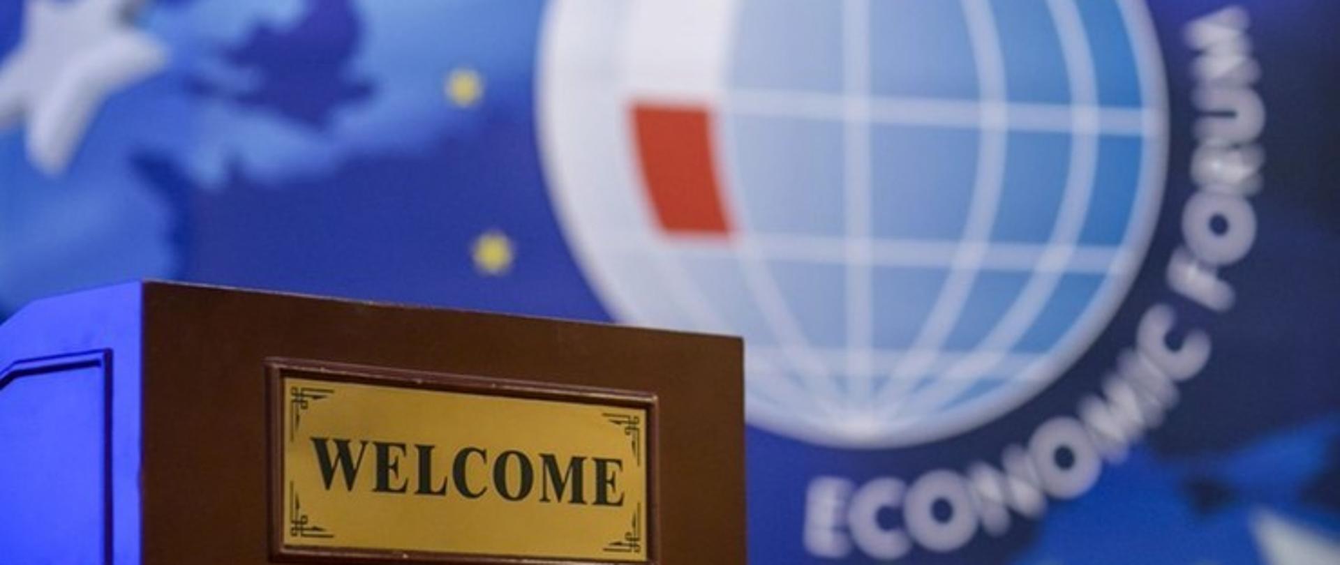 na tle logo Forum Ekonomicznego - kula ziemska z białoczerwonymi elementami symbolizującymi flagę Polski. wokół flagi napis Economic Forum. statyw mównicy z napisem WELCOME