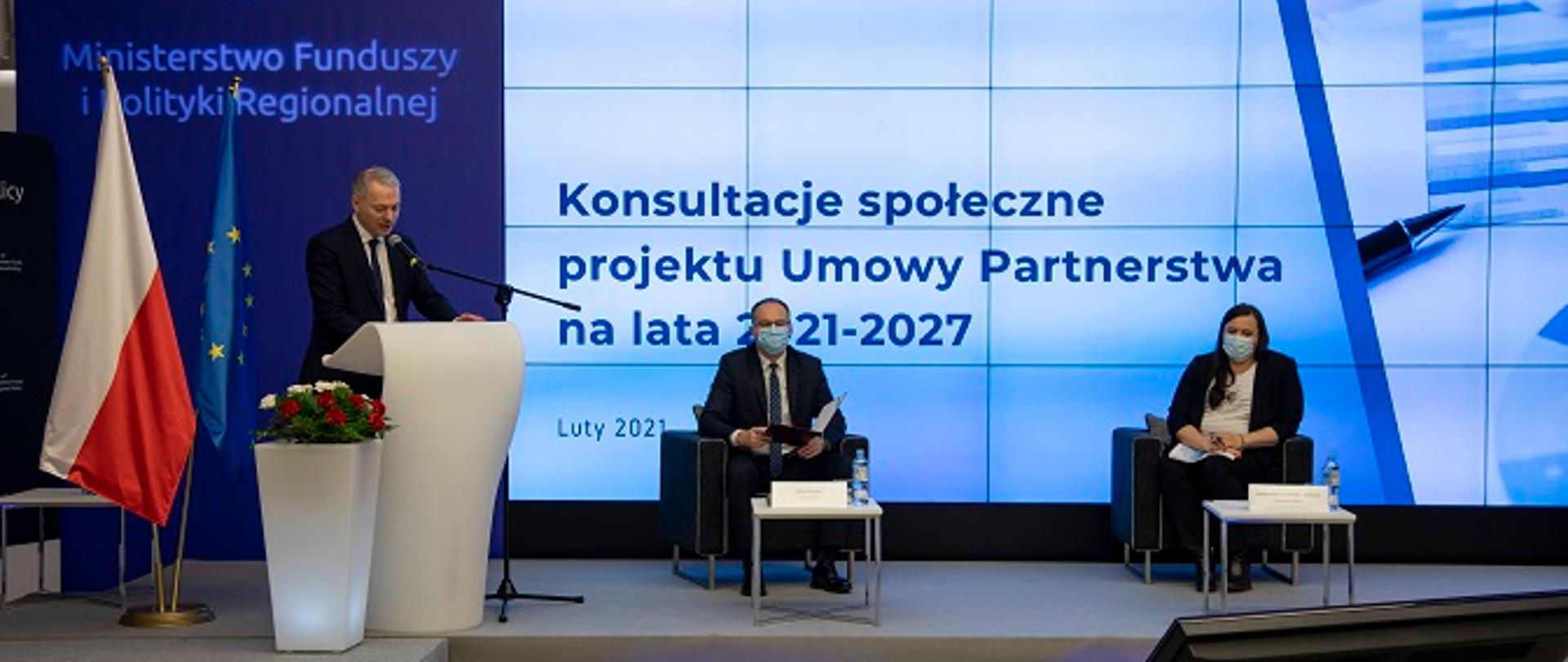 Zdjęcie przedstawia konsultacje Umowy Partnerstwa w woj. podlaskim, wiceminister Jacek Żalek przemawia przy mównicy, obok siedzą prowadzący konferencję Grzegorz Osiecki oraz wiceminister Małgorzata Jarosińska-Jedynak. W tle flagi Polski i UE oraz ekran z napisem Konsultacje społeczne projektu Umowy Partnerstwa na lata 2021-2027