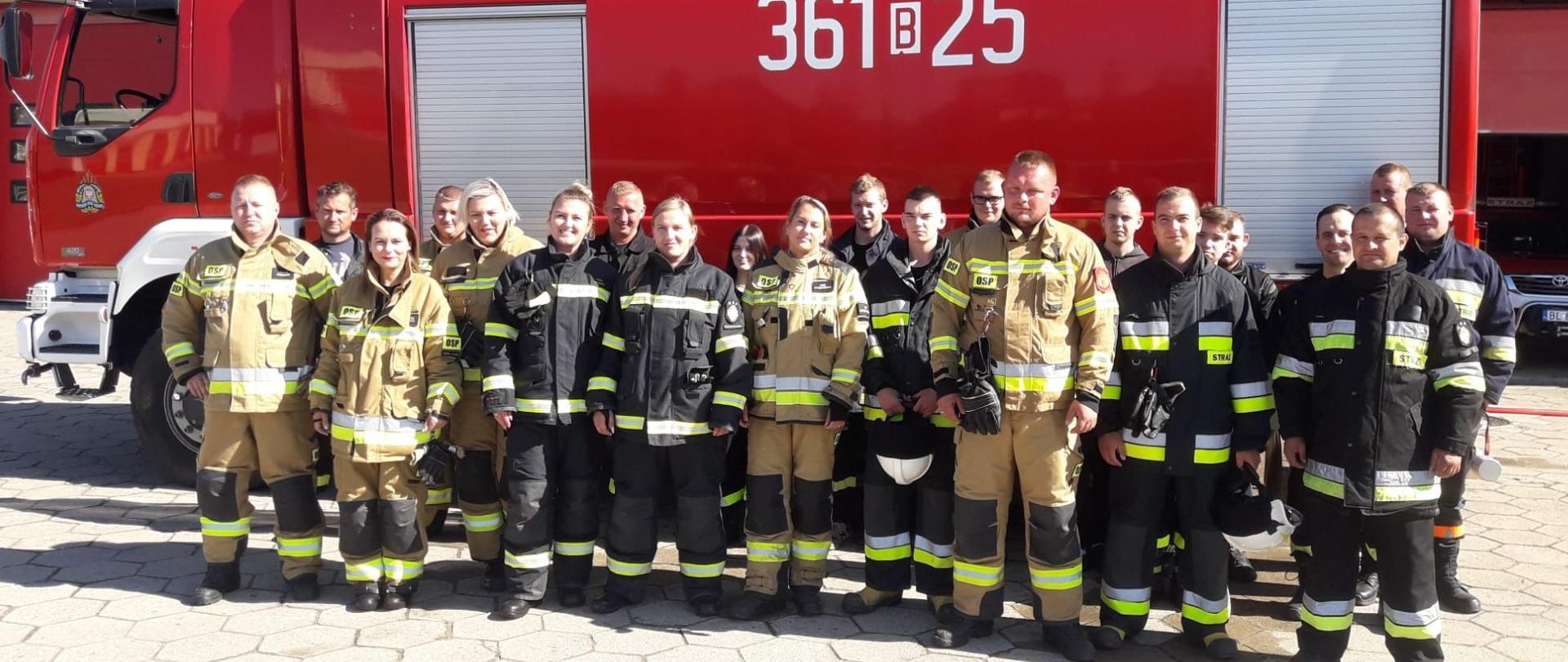 Zdjęcie grupowe uczestników szkolenia podstawowego OSP. Wszyscy strażacy ubrani w umundurowanie specjalne. W tle widoczny samochód pożarniczy.
