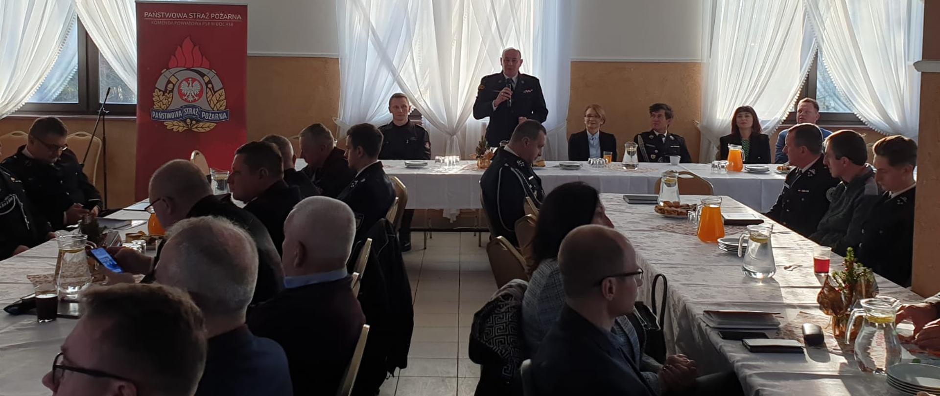 Zdjęcie przedstawia kilkudziesięciu strażaków ubranych w ubrania koszarowe, którzy uczestniczą w naradzie operacyjno-szkoleniowej. Siedzą przy stołach . W tle strażak ubrany w umundurowanie wyjściowe stoi i trzyma w ręku mikrofon i przemawia. W tle widać zasłony w oknach koloru białego