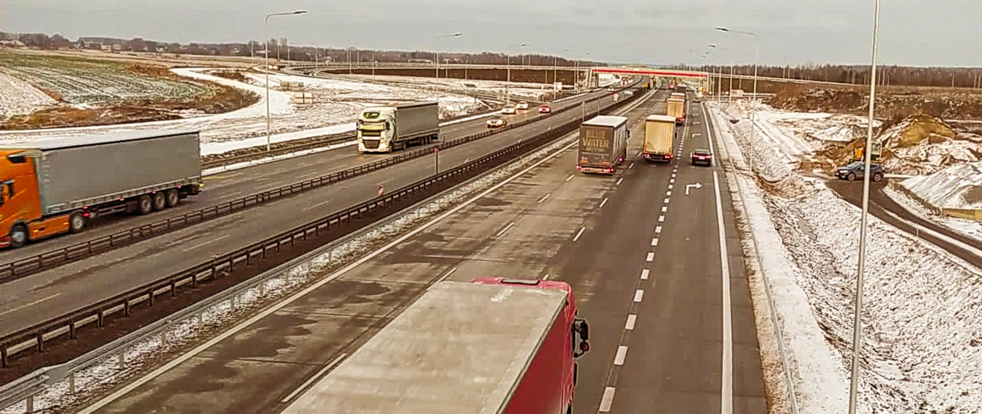Fotografia dwujezdniowej autostrady A1 z poziomu obiektu inżynierskiego. Na fotografii widoczne jezdnie, po których odbywa się dwukierunkowo ruch. W tle widoczny obiekt mostowy przecinający poprzecznie autostradę. Na obrzeżach fotografii widoczne prace ziemne w aurze zimowe, pobocza pokryte śniegiem.
