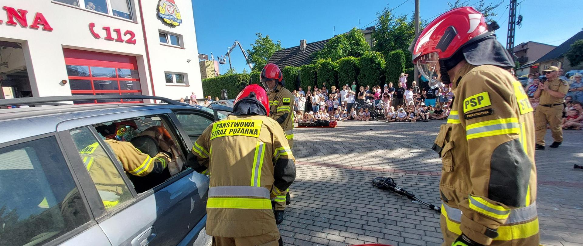 Zdjęcie przedstawia pokaz strażacki na świeżym powietrzu, prowadzony prawdopodobnie dla uczestników pikniku strażackiego. Na pierwszym planie trzej strażacy w pełnym rynsztunku ochronnym – piaskowych kombinezonach z żółtymi odblaskowymi pasami i czerwonych hełmach – aktywnie działają wokół srebrnego samochodu. Jeden strażak wychylał się do środka przez otwarte okno od strony pasażera, a drugi stał w pobliżu, zwrócony twarzą do samochodu. Trzeci strażak był po prawej stronie, zwrócony twarzą do samochodu, z napisem „PSP” (Państwowa Straż Pożarna) widocznym na hełmie i spodniach. Na utwardzonym podłożu w pobliżu leżały różne narzędzia, w tym coś, co wyglądało na hydrauliczny sprzęt ratowniczy. W tle widać tłum ludzi, w tym wiele dzieci i młodzieży, obserwujących demonstrację. Za nimi widać biały budynek z czerwonymi akcentami, który prawdopodobnie jest budynkiem komendy PSP w Przysusze (oznaczoną numerem „112” i napisem „Państwowa Straż Pożarna” po polsku). Akcja rozgrywa się na świeżym powietrzu, w jasny, słoneczny dzień.