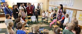 Odwiedziny w krapkowickiej szkole. Na zdjęciu widać dzieci. Każde z nich stoi za swoim krzesłem. Krzesła ułożone są dookoła, tworząc okrąg. Po środku widać walizkę z maskotką, która jest elementem ich zabawy. W tle widać uczestników odwiedzin w krapkowickiej szkole, między innymi Wiceminister Katarzynę Lubnauer. 