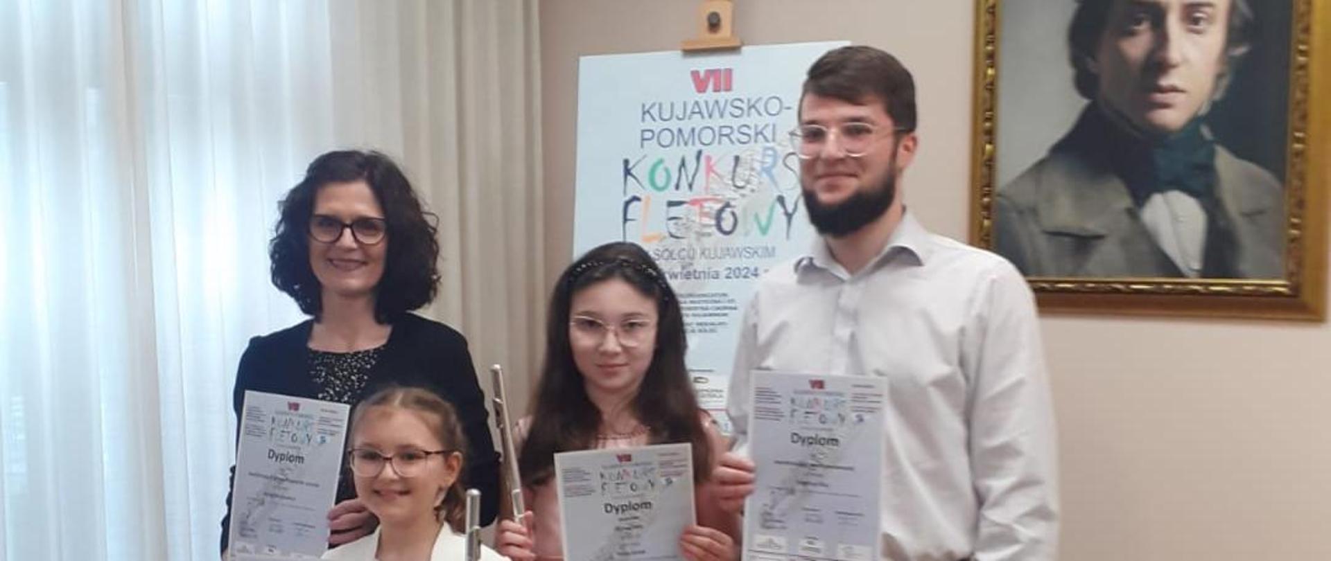 Od lewej Pani Anna Wojtowicz, Natalia Paliwoda, Maja Solak i pan Krystian Klej. Osoby trzymają dyplomy. W tle na sztaludze plakat konkursu. Na ścianie obraz kompozytora.