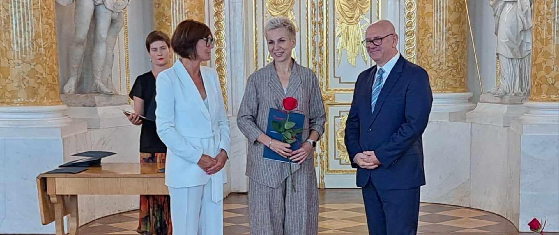 Wnętrze sali w Zamku Królewskim w Warszawie, na pierwszym planie stoją: po środku pani dyrektor Małgorzata Wagner po jej lewej i prawej stronie wręczający nominację.