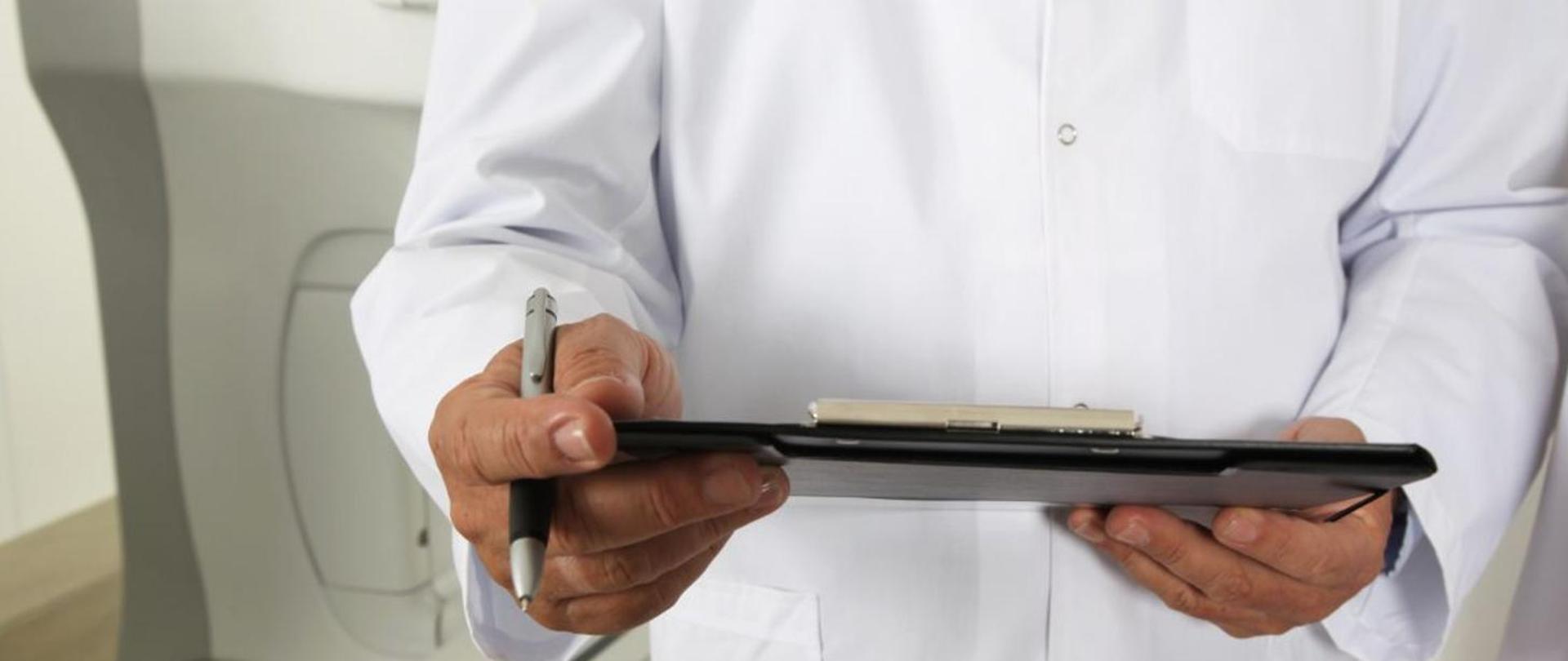 Zdjęcie przedstawia pracownika medycznego, ubranego na biało, trzymającego w ręku tablet