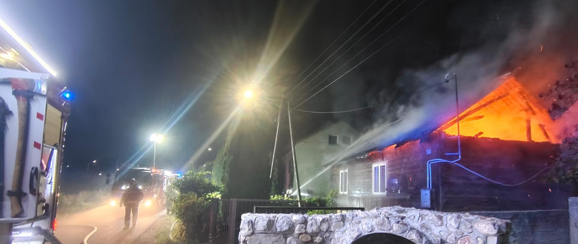 Po prawej stronie widać budynek drewniany, którego poddasze płonie, z lewej strony widać wozy strażackie i idącego drogą ratownika.