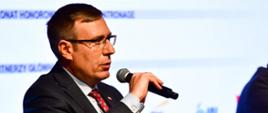 Wiceminister Maciej Małecki przemawia z fotelika. W tle ekran z logo konferencji. Obok siedzą pozostali paneliści.