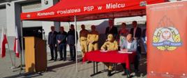 Fotografia wykonana na placu komendy powiatowej Państwowej Straży Pożarnej w słoneczny dzień podczas uroczystości przekazania placu wykonawcy pod rozbudowę i modernizację komendy powiatowej i jednostki ratowniczo – gaśniczej nr 1. Zdjęcie wykonane w momencie podpisywania protokołów przekazania i przejęcia placu pod rozbudowę odpowiednio przez komendanta powiatowego i wykonawcę inwestycji. Osoby siedzą przy stoliku podpisując dokumenty. Za nimi stoją zaproszeni goście. Wszyscy stoją pod czerwonym namiotem z napisem komenda powiatowa PSP w Mielcu. Na pierwszym planie widać baner komendy powiatowej z logo Państwowej Straży Pożarnej.