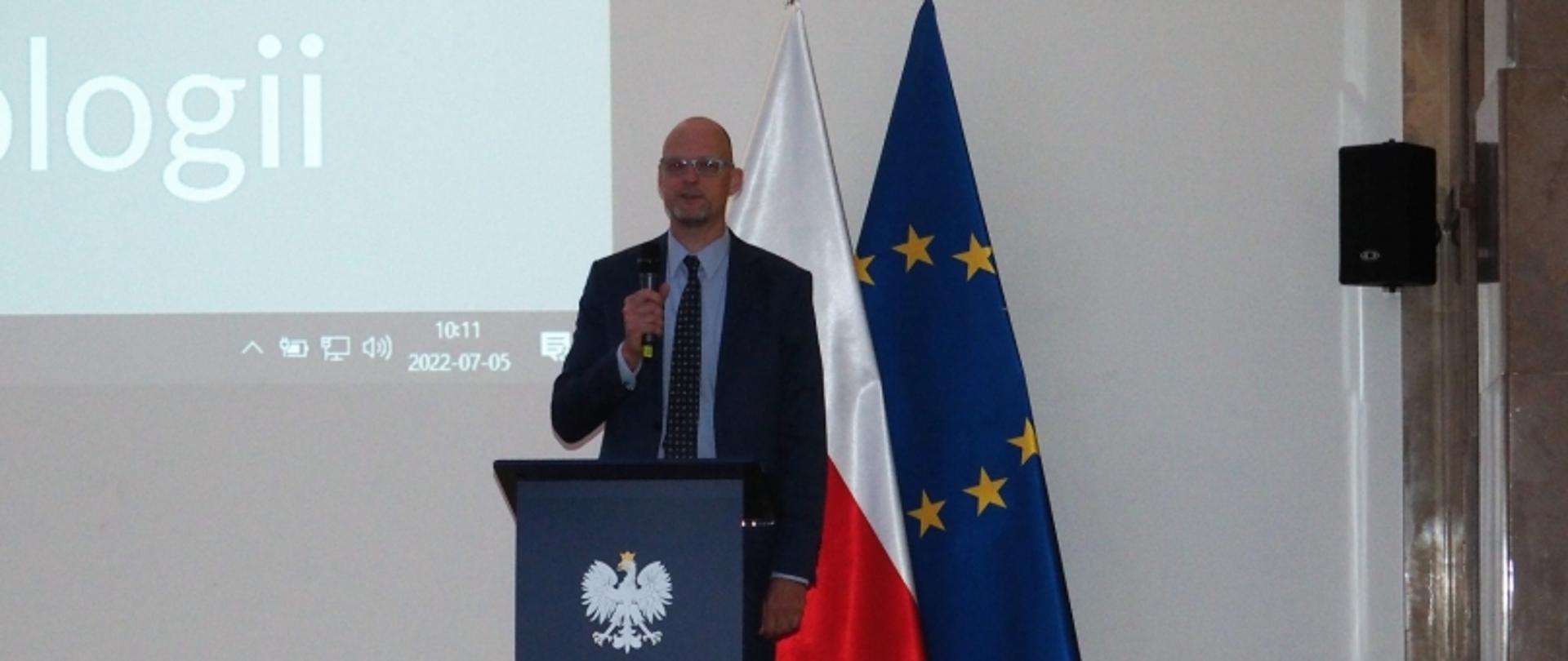 Osoba przy mównicy z godłem Polski, w tle flagi Polski i UE oraz ekran z napisem „technologii” i datą.