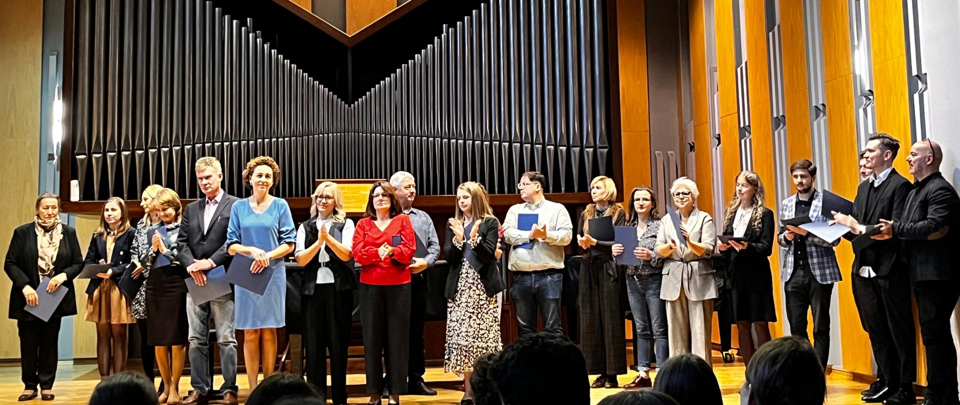 Zdjęcie przedstawia dużą grupę osób - nauczycieli stojących na scenie w sali koncertowej podczas wręczenie nagród.
W tle dominuje monumentalny, nowoczesny prospekt organowy w ciemnych barwach, którego piszczałki ułożone są symetrycznie w kształt litery "V" lub odwróconej góry. Piszczałki są lśniące, co sugeruje metal. Nad prospektem w suficie znajdują się trzy rzędy jasnych, okrągłych świateł punktowych.
Po prawej stronie sceny widoczna jest ściana z pionowymi panelami, część z nich jest drewniana lub w kolorze jasnego drewna, a część ciemnoszara, nadając wnętrzu współczesny, elegancki charakter.