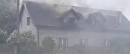 Zdjęcie przedstawia pożar budynku mieszkalnego wielorodzinnego. Dym unosi się nad dachem budynku. 