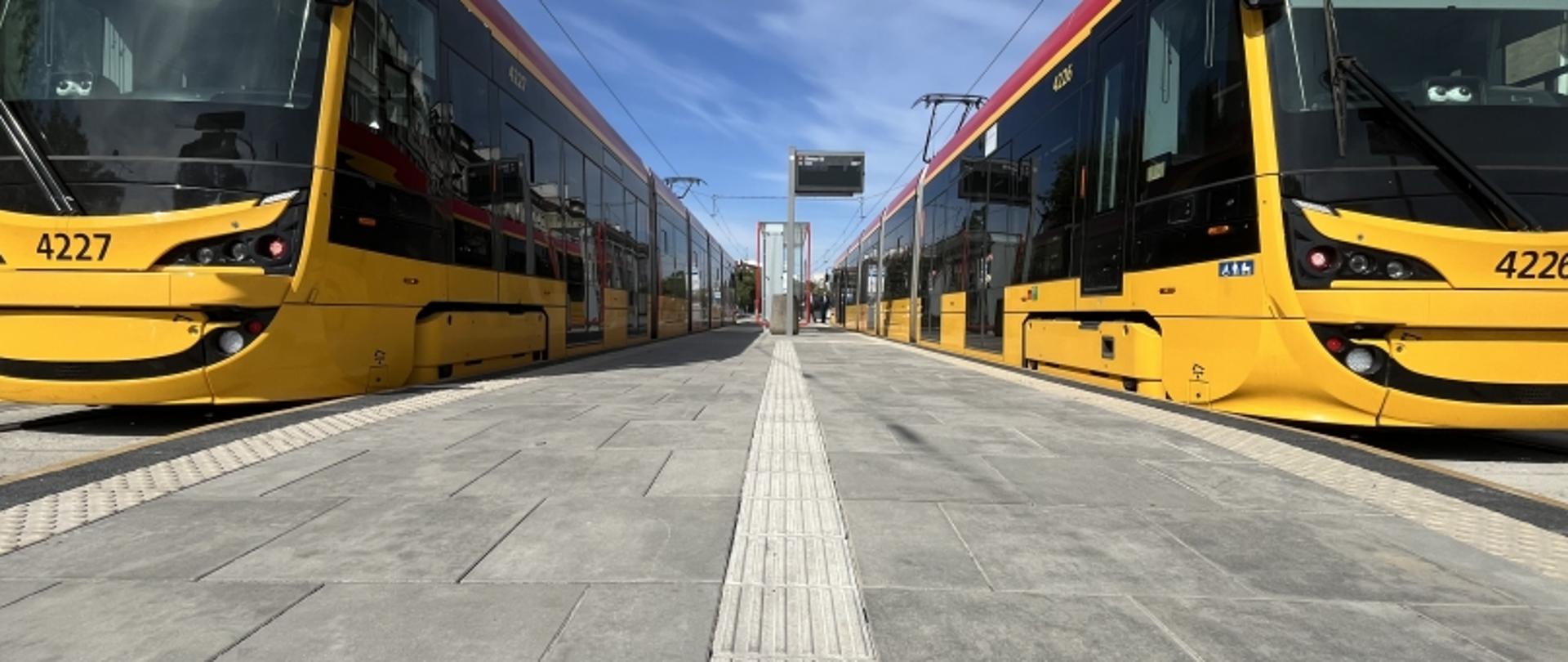 Dwa żółte tramwaje naprzeciw siebie na torach, z peronem pośrodku i niebieskim niebem w tle.
