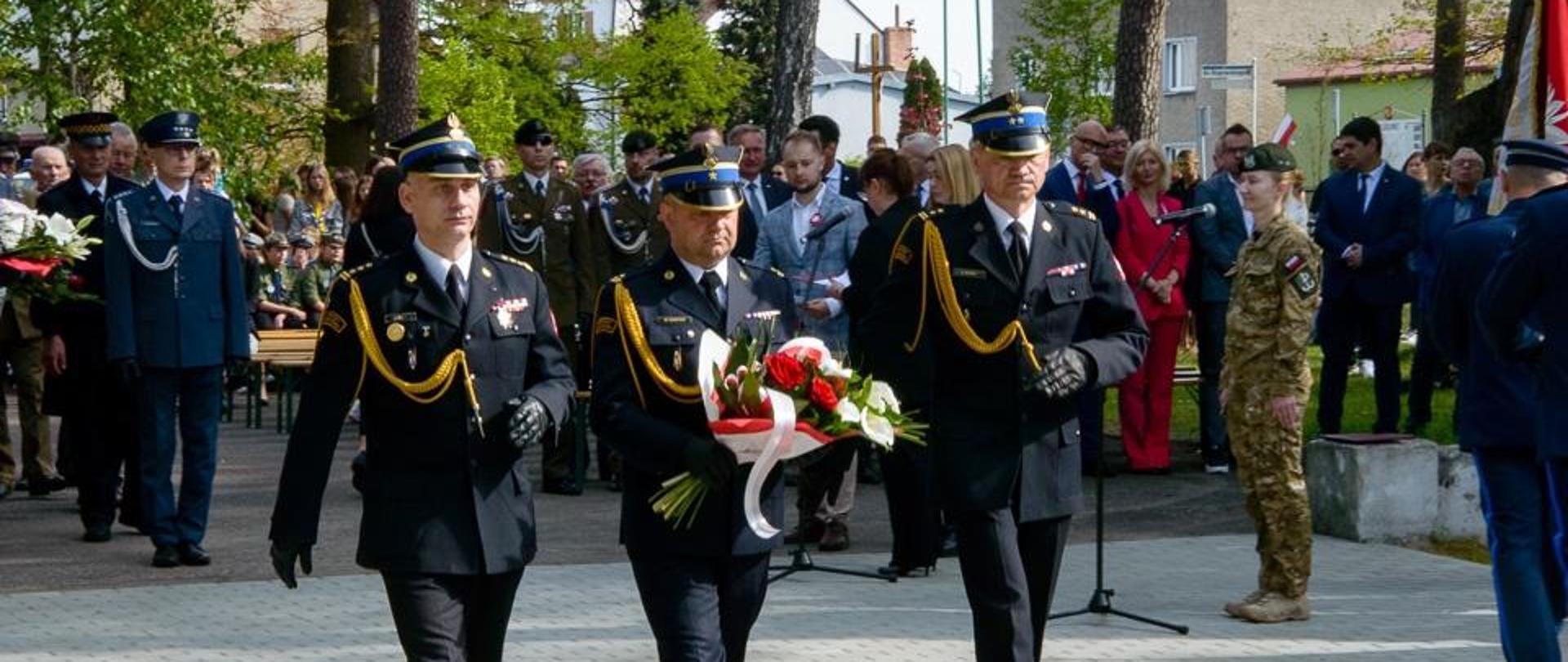 Na tle zaproszonych gości i delegacji, delegacja trzech funkcjonariuszy Państwowej Straży Pożarnej w umundurowaniu galowym, jeden z nich trzyma wieniec.