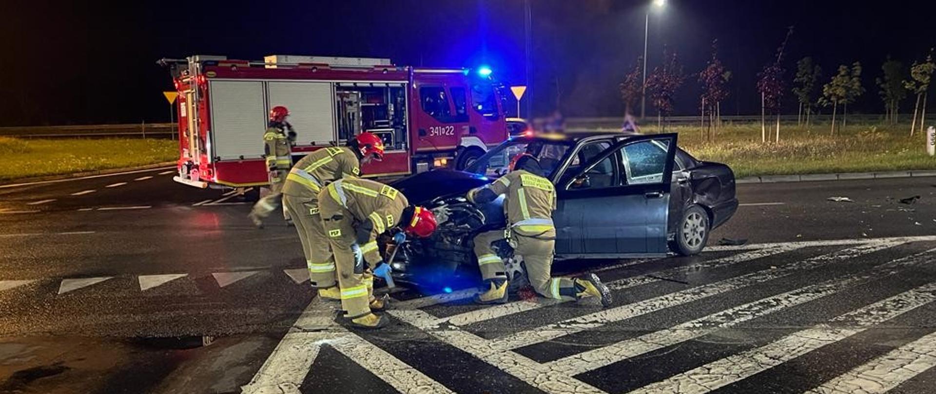 Strażacy wykonują czynności [przy samochodzie osobowym. W tle samochód pożarniczy.