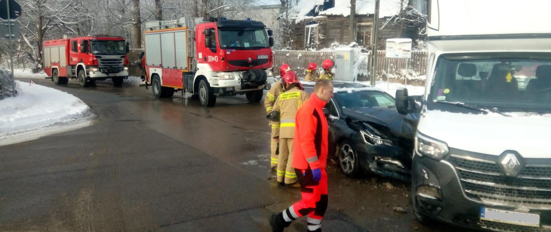 Po prawej stronie na pierwszym planie stoi laweta biorąca udział w zderzeniu. Przy niej z lewej strony samochód osobowy – widać znaczne uszkodzenia przodu auta. Przy zniszczonych pojazdach ratownik medyczny i dwóch strażaków. W głębi ulicy stoją zaparkowane dwa samochody pożarnicze. 