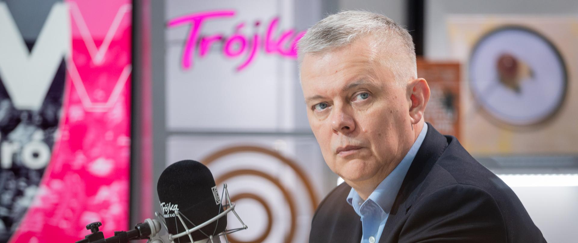 Na zdjęciu studio radiowe. Na pierwszym planie po prawej stronie za mikrofonem siedzi minister Tomasz Siemoniak. W lewym dolnym rogu widać fragment stołu. Po lewej stronie zdjęcia widać mikrofon radiowy z napisem "Trójka Polskie Radio". W lewym górnym rogu fragment ekranu z różowo-czarnym tłem. W tle widać różowy neon "Trójka" oraz inne elementy dekoracji studia m.in. płyty.