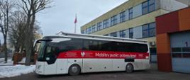 Zdjęcie przedstawia duży, biały autokar zaparkowany na placu przed budynkiem komendy straży pożarnej. Na boku pojazdu widnieje szeroki czerwony pas z napisem „Ministerstwo Zdrowia – Mobilny punkt poboru krwi” oraz logo Narodowego Centrum Krwi. W tle widać budynek z niebieskimi ramami okiennymi oraz wjazd do garażu strażackiego z numerem 1.