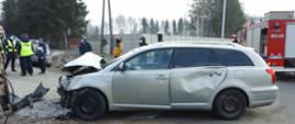 Srebrny samochód osobowy po zderzeniu z samochodem dostawczym. Przód pojazdu jest rozbity, uszkodzone tylne drzwi od strony kierowcy. W tle wóz strażacki, strażacy, policjanci oraz osoby postronne.
