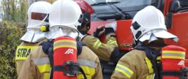 Ćwiczenia w ZOL. Zdjęcie przedstawia 4 strażaków na tle fragmentu wozu strażackiego. Wszyscy mają na sobie ubrania specjalne koloru piaskowego z biało-żółtymi pasami odblaskowymi Trzech z nich ma hełmy w kolorze białym, czwarty - w czerwonym. Dwóch strażaków wyposażonych jest w aparaty oddechowe, których butle znajdują się w czerwonych pokrowcach. Strażak w czerwonym hełmie, na masce samochodu, odnotowuje dane w karcie kontrolnej. W tle krzewy i drzewa. 