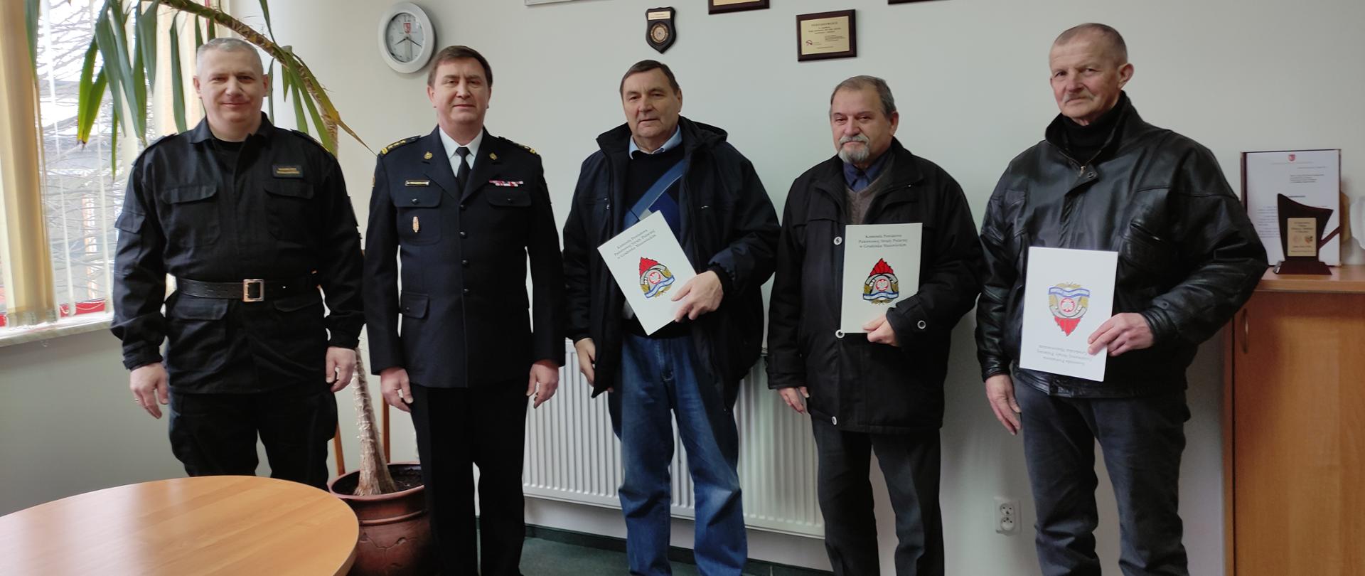 Wspolnie stoją st. bryg. Krzysztof Tryniszewski - Komendant Powiatowy PSP w Grodzicku Mazowieckim, kpt. Mariusz Wołowski - Dowódca JRG oraz druhowie, którym wręczono decyzje o przyznaniu świadczenia ratowniczego.